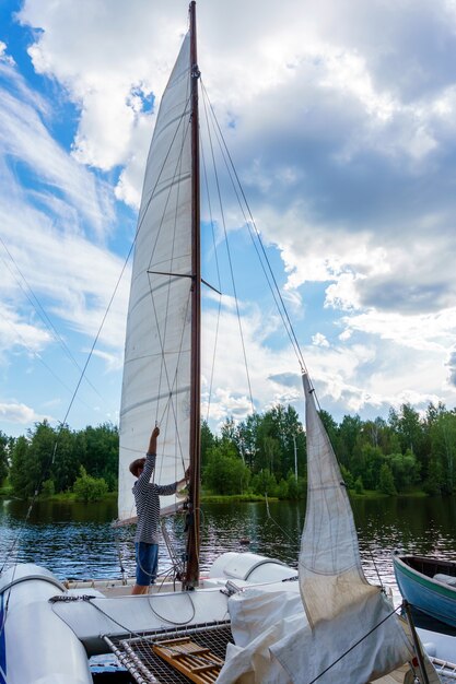 Photo young sailor raises a sail on a small sailing catamaran moored in a wooded bay