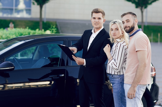 A young salesman shows a new car to customers. Man and woman buy a car.