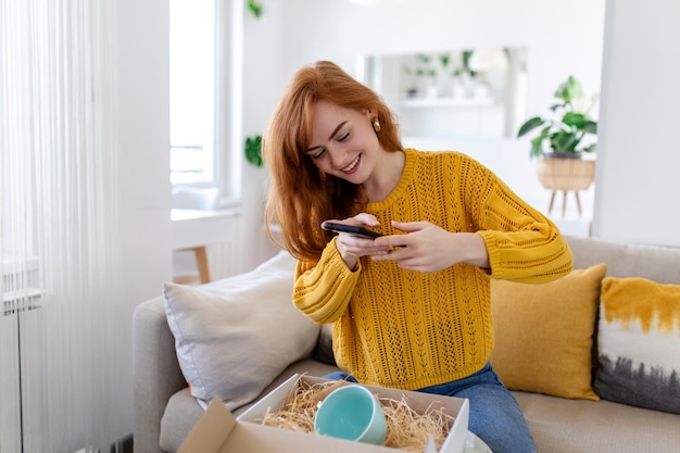 Young satisfied happy woman shopaholic customer sit on sofa unpack parcel delivery box online shopping shipment concept taking photos of product to post on social media