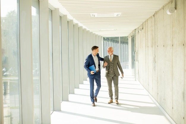 Young and a senior businessman walk down an office hallway deep in conversation