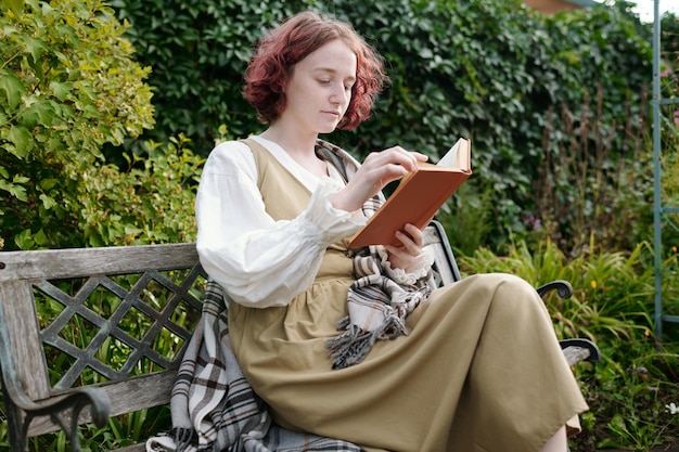 Young serene woman in casual dress sitting on bench in the garden and reading