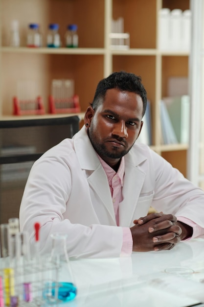 Young serious african american scientist in lab coat looking at camera while sitting by desk with te