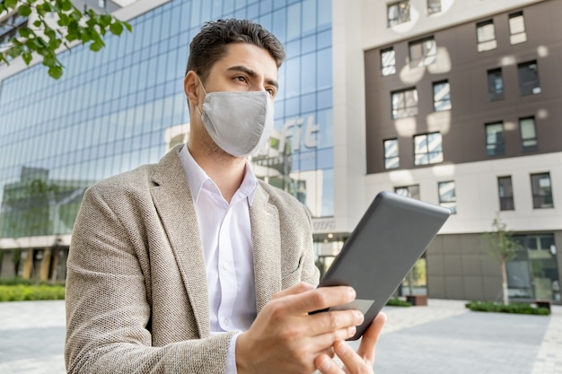 Young serious businessman in formalwear and protective mask using touchpad while standing against modern office building in park