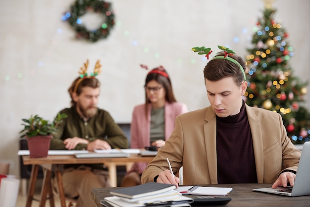 Young serious businessman in xmas headwear making notes in notebook while planning work by desk in front of laptop on Christmas eve