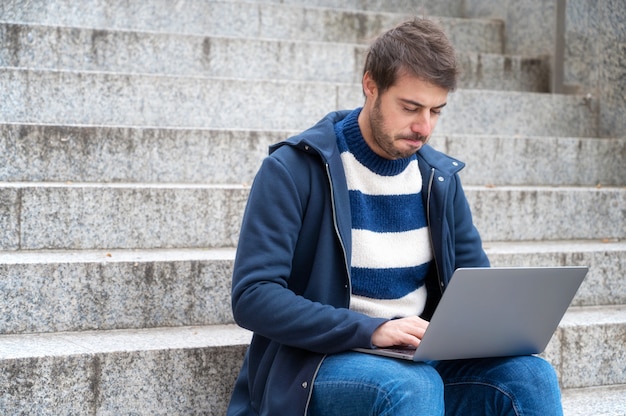 young serious expression, man freelancer working with laptop outdoors sitting at stair steps, in urban scenery.