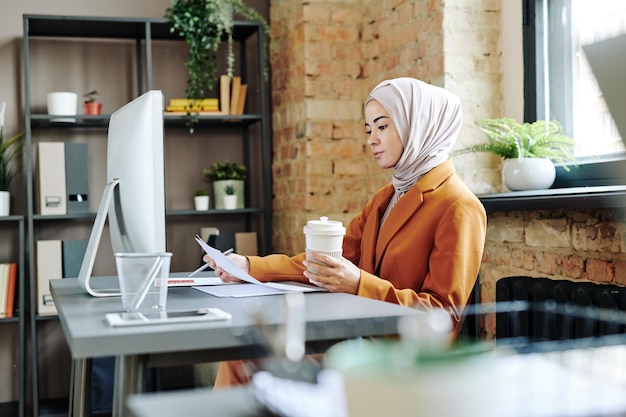 Young serious female accountant in hijab looking through financial papers