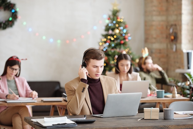 Young serious male customer support representative with headset looking at laptop display while sitting by desk and consulting clients