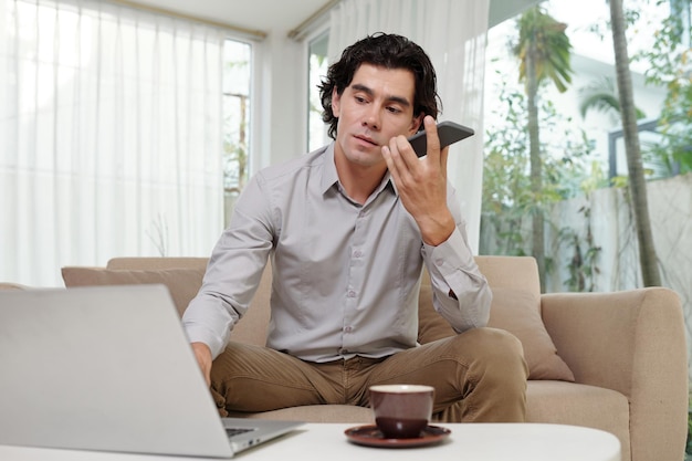 Young serious male employee talking by smartphone and using laptop