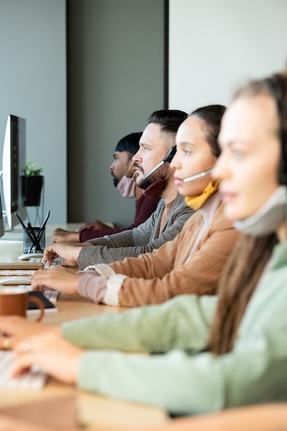 Young serious male operator and his colleagues with protective masks under chins consulting clients online while working in call center