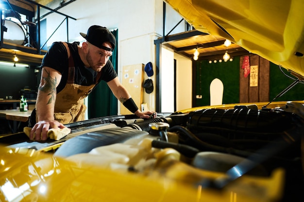 Young serious man in apron and baseball cap bending over open hood of car