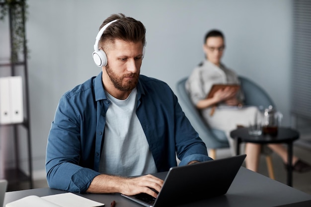 Young serious man in headphones and casualwear sitting in front of laptop