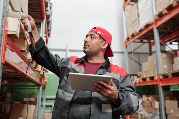 Young serious mixed-race manager of warehouse with touchpad fixing prices in electronic document while scanning qr codes on boxes