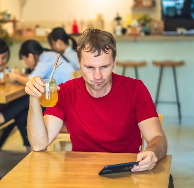 Young serious thoughtful pensive attentive man tense thinking send message by smart phone in red shirt in cafe drinking chia ice tea Real people expression life problems solving and communication