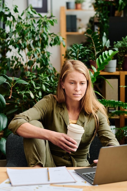 Young serious woman with cup of coffee sitting by workplace in front of laptop