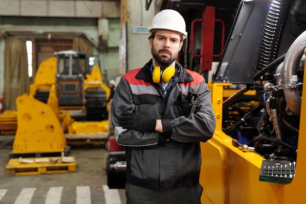 Young serious worker of plant in hardhat and uniform standing by huge machine