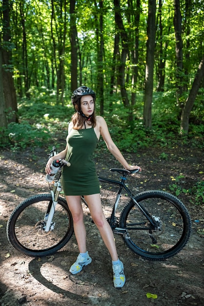 Young sexy active woman in green dress stand near bicycle in the forest Activity lifestyle in summer day