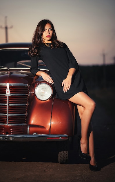 Young sexy brunette with long legs in a black dress and high-heeled shoes lies on the hood of a burgundy retro car on a country road in the late evening