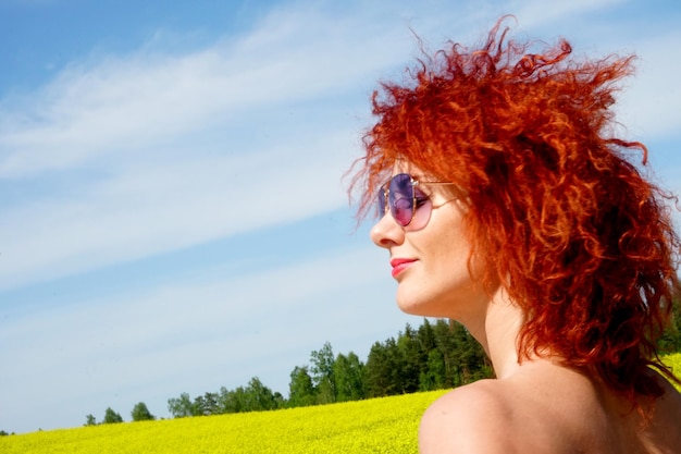 Young sexy Caucasian woman with red hair and sunglasses on yellow rape field background, summer day