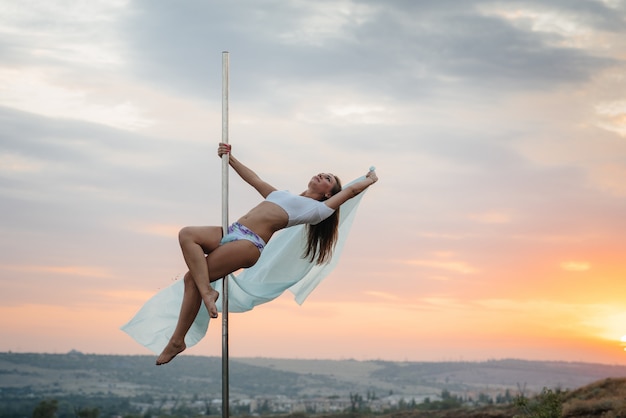 A young sexy girl performs amazing exercises on a pole during a beautiful sunset.