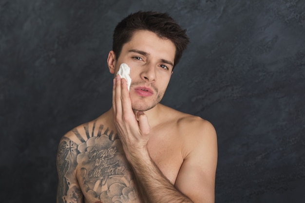 Young sexy tattooed man put foam on face. Topless guy having fun while shaving, gray studio background