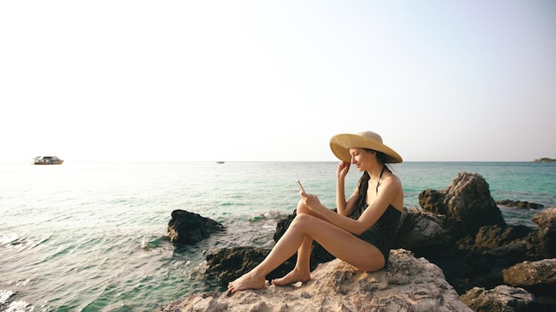 Young sexy woman in swimsuit and hat making selfie shoot using smartphone during vacation on beach