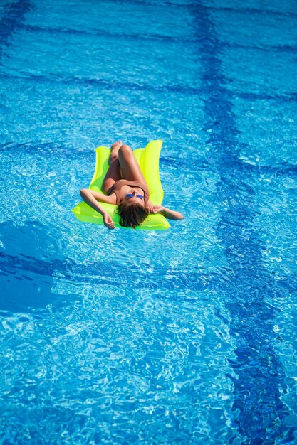 Young sexy woman in a swimsuit swims in the pool on an inflatable mattress. Girl in sunglasses in a blue pool on vacation