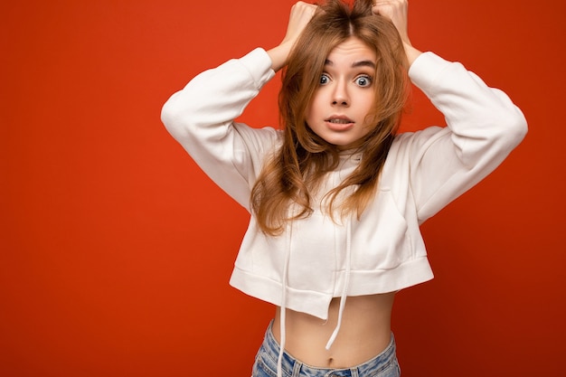 Young shocked amazed beautiful dark blond woman with sincere emotions isolated on wall wall with copy space wearing casual white hoodie. Surprise concept.