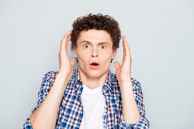 Young shocked guy raising hands to head isolated over grey