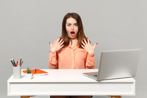 Young shocked woman in pastel clothes keeping mouth open, spreading hands work at desk with pc laptop isolated on gray background. Achievement business career lifestyle concept. Mock up copy space.