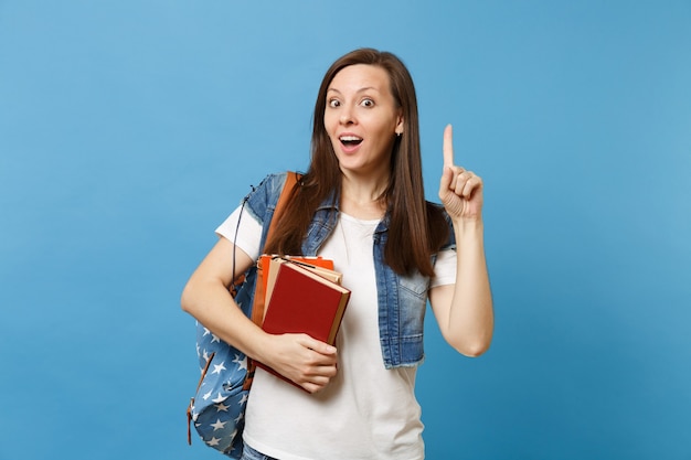 Young shocked woman student with backpack pointing index finger up on copy space and got new idea holding school books isolated on blue background. Education in high school university college concept.