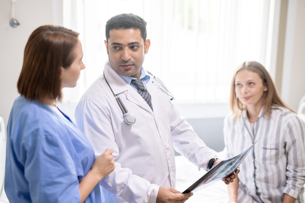 Young sick woman sitting on bed and looking at confident doctor discussing x-ray results with his assistant in hospital chamber