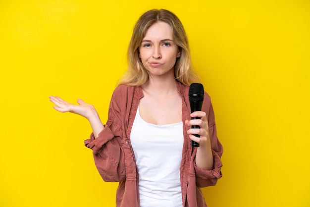 Young singer caucasian woman picking up a microphone isolated on yellow background having doubts while raising hands