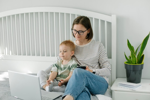 Young single mother mom with toddler boy son sitting on bed using laptop searching web watching cartoons