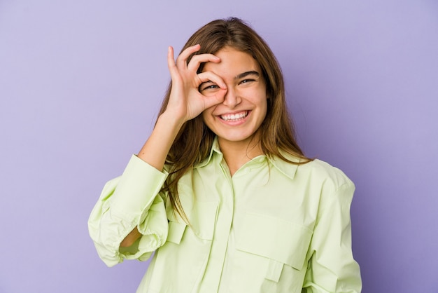 Young skinny caucasian girl teenager on purple background excited keeping ok gesture on eye.