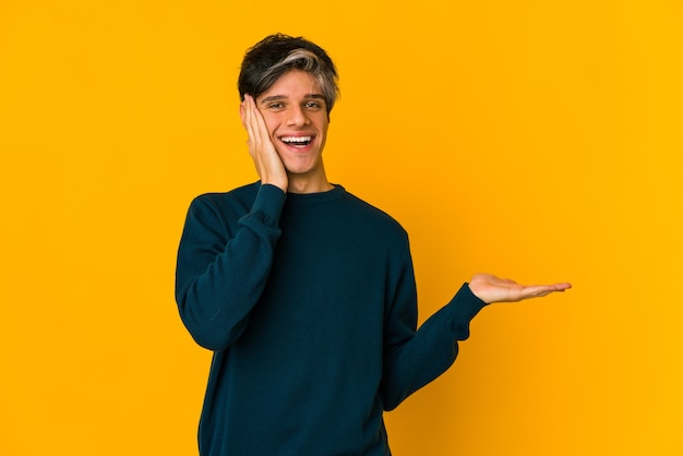 Young skinny hispanic man holds copy space on a palm, keep hand over cheek. Amazed and delighted.