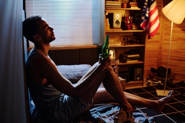 Young sleepless man with bottle of alcohol sitting on bed against wall