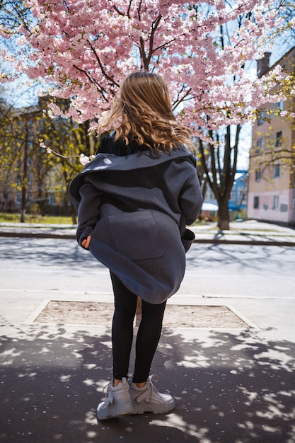 Young slender female model with long wavy hair and, dressed in a gray coat, sneakers, spinning on the street. Spring blossom trees woman girl laughs and runs to rejoice. Selective focus