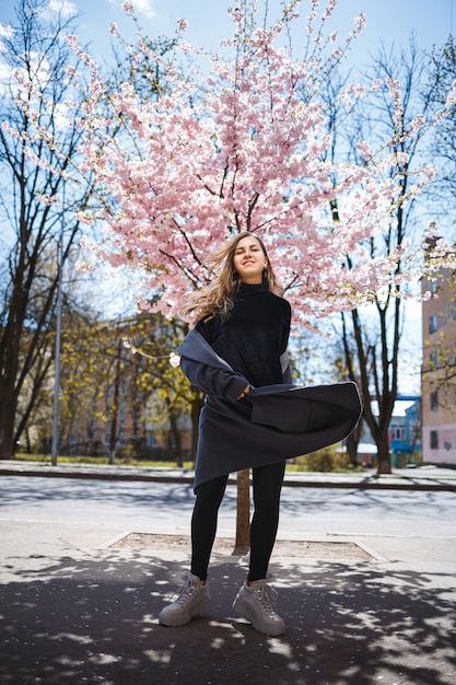 Young slender female model with long wavy hair and, dressed in a gray coat, sneakers, spinning on the street. Spring blossom trees woman girl laughs and runs to rejoice