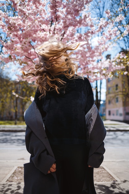 Young slender female model with long wavy hair and, dressed in a gray coat, sneakers, spinning on the street. Spring blossom trees woman girl laughs and runs to rejoice