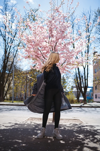 Young slender female model with long wavy hair and, dressed in a gray coat, sneakers, spinning on the street. Spring blossom trees woman girl laughs and runs to rejoice