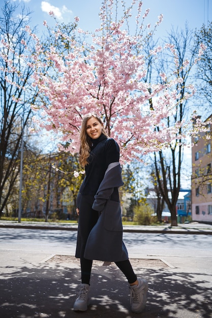 Young slender female model with long wavy hair and, dressed in a gray coat, sneakers, spinning on the street. Spring blossom trees woman girl laughs and runs to rejoice