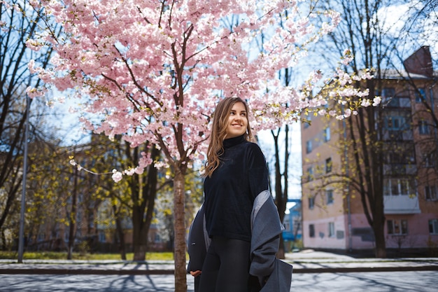 A young slender female model with long wavy hair and, dressed in a gray coat, sneakers, stands on the street near a flowering shrub with beautiful pink flowers in the background and poses.