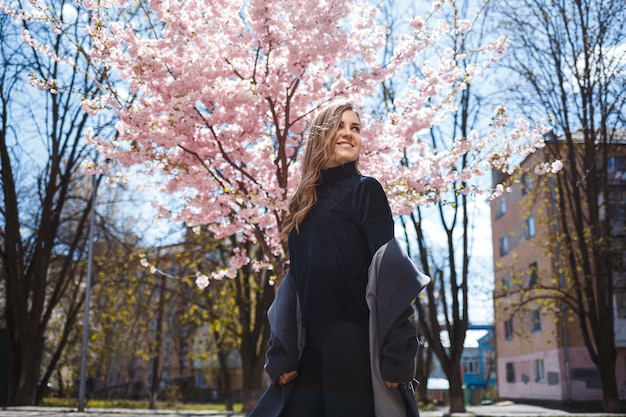 A young slender female model with long wavy hair and, dressed in a gray coat, sneakers, stands on the street near a flowering shrub with beautiful pink flowers in the background and poses.