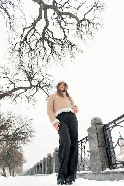 Young slender girl in black trousers and beige sheepskin coat in snowcovered park Stylish woman Vertical frame