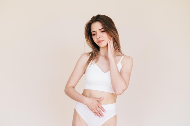 Young Slender Gloomy Woman in White Underwear Hand on Stomach on Beige Background