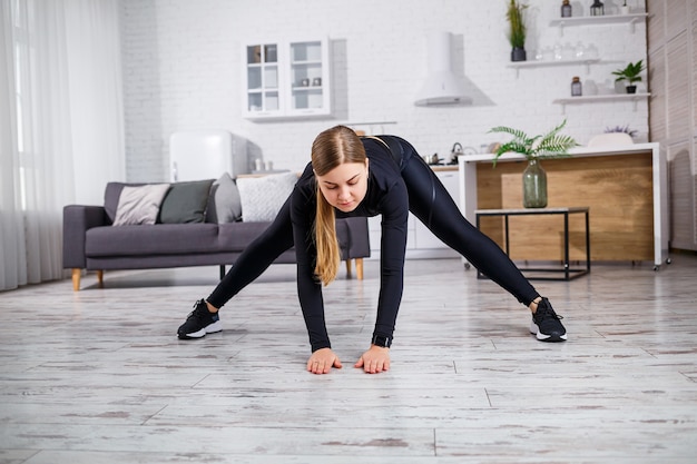 A young slender woman goes in for sports at home, wearing fitness clothes, a black top and leggings. Fitness at home for a beautiful body. Warm up before playing sports