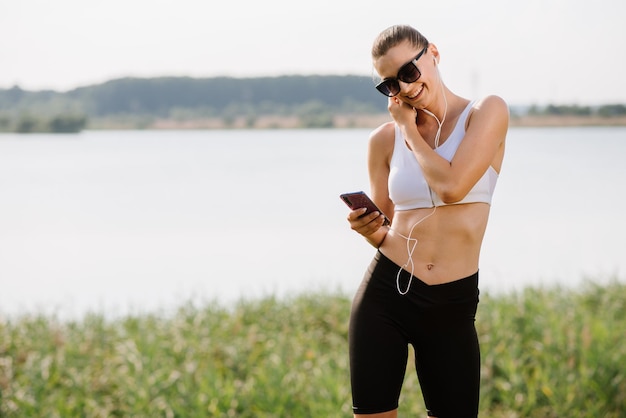 Young slim fit woman with earphones and smartphone listen music in the park at the sunny day