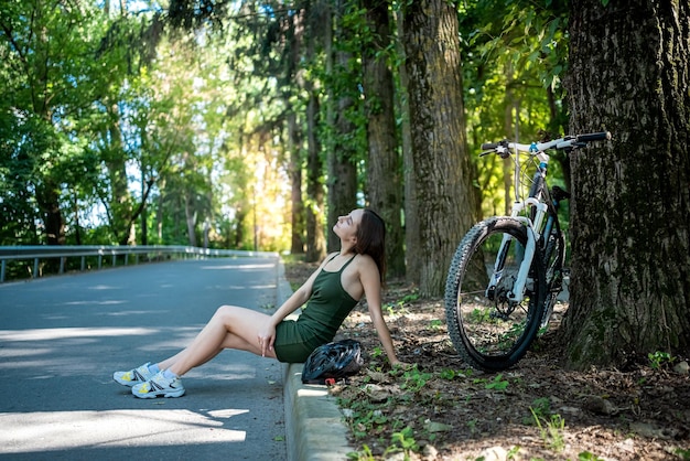 Young slim woman in green dress on bike standing on road and rest after trip