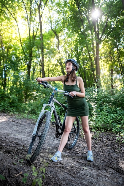 Young slim woman in green dress and hardhat for safety stand near bike on park