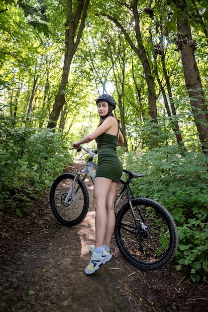 Young slim woman in green dress and hardhat for safety stand near bike on park